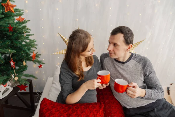 Pareja familiar en la cama en la habitación de Año Nuevo sosteniendo tazas rojas con té . —  Fotos de Stock