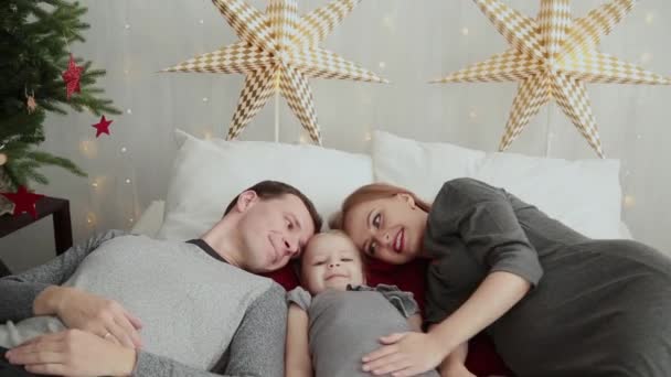 Manhã de Natal. A jovem família brinca alegremente uns com os outros. Em cima da cama um monte de almofadas, presentes de Natal em embalagens brilhantes e decorações de Natal. Feliz Natal 2019 . — Vídeo de Stock