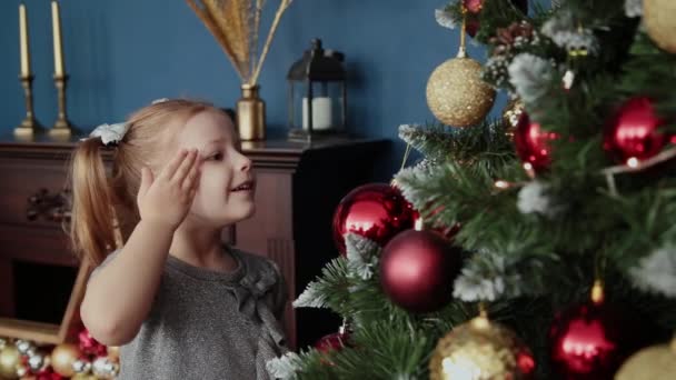 Velmi krásná holčička stojí poblíž stromu nový rok a dotkne hračky. — Stock video