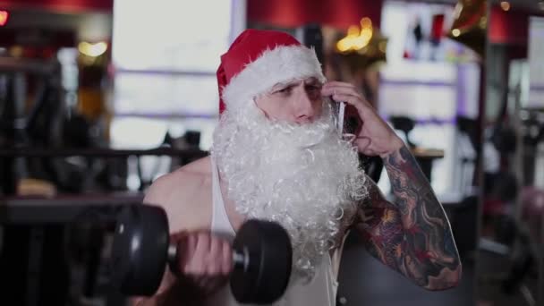 Instructor de fitness Santa Claus en el gimnasio levantando una mancuerna y hablando por teléfono . — Vídeos de Stock