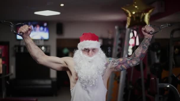 Instruktor fitness Santa Claus w siłowni pociągi rękami na symulatorze i grymasy. — Wideo stockowe