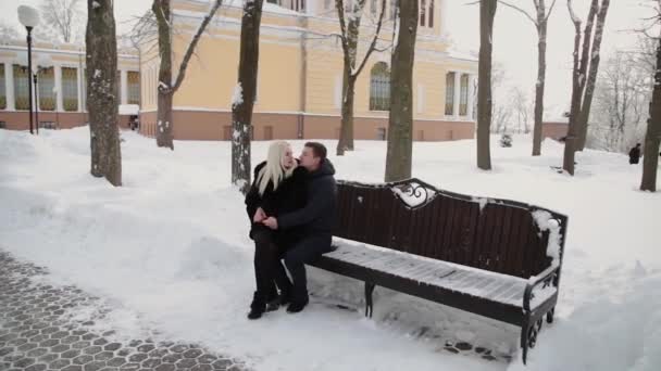 Hermosa pareja joven sentada en un banco abrazo hablando y sonriendo en un parque de invierno de la ciudad . — Vídeos de Stock