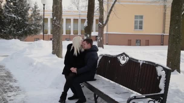 Όμορφο νεαρό ζευγάρι κάθεται σε ένα παγκάκι αγκαλιάζω μιλάμε και χαμογελαστά σε ένα χειμερινό πάρκο της πόλης. — Αρχείο Βίντεο