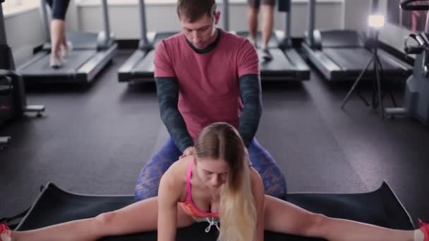 Atletisk och attraktiv man hjälper en vacker kvinna att sträcka på golvet i gymmet. — Stockvideo