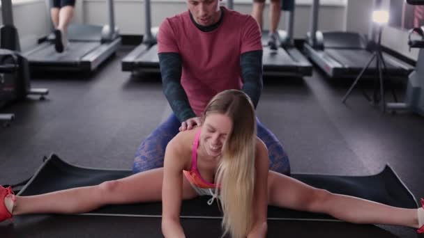 Atlético y atractivo hombre ayuda a una hermosa mujer a estirarse en el suelo en el gimnasio . — Vídeo de stock