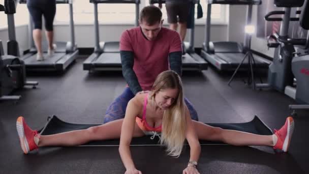 Homem atlético e atraente ajuda uma mulher bonita a se esticar no chão no ginásio . — Vídeo de Stock