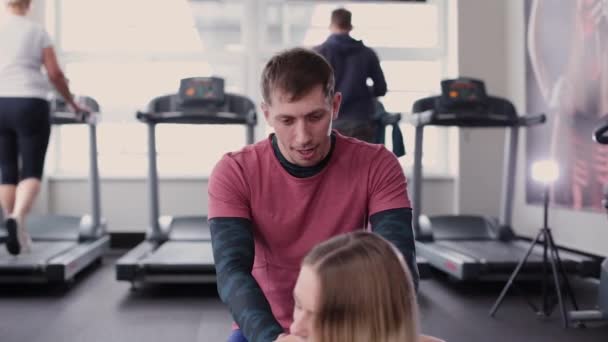 Athletic and attractive man helps a beautiful woman to stretch on the floor in the gym. — Stock Video