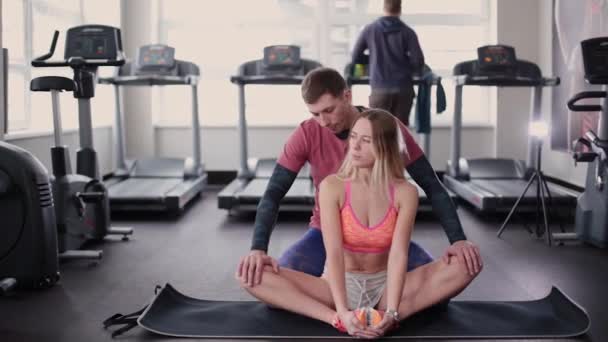 Atletisk och attraktiv man hjälper en vacker kvinna att sträcka på golvet i gymmet. — Stockvideo