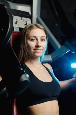 Aktif güzel fitness modeli kız göğüs kasları bir simülatör spor salonunda trenler.