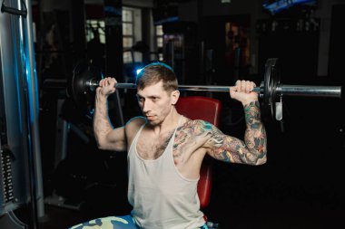 Yakışıklı atletik spor salonunda bankta oturmuş halter kaldırma omuz trenler.