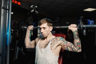 Yakışıklı atletik spor salonunda bankta oturmuş halter kaldırma omuz trenler.