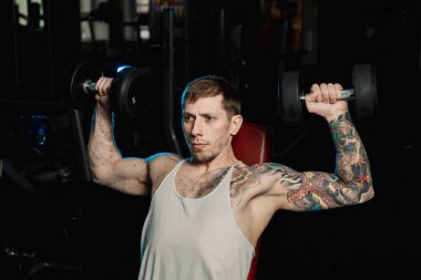 Spor salonunda bir bankta oturmuş dumbbells ile omuzlarını eğitim yakışıklı atletik erkek.