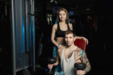 Aktif güzel fitness modeli kız erkek spor salonunda otururken dumbbells ile tren yardımcı olur.