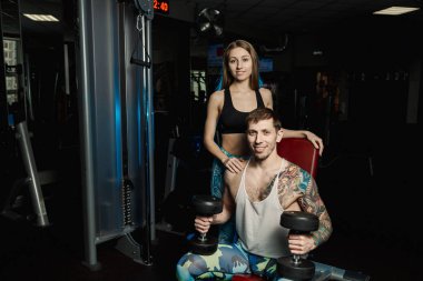 Aktif güzel fitness modeli kız erkek spor salonunda otururken dumbbells ile tren yardımcı olur.