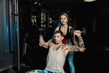 Aktif güzel fitness modeli kız erkek spor salonunda otururken dumbbells ile tren yardımcı olur.