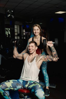 Aktif güzel fitness modeli kız erkek spor salonunda otururken dumbbells ile tren yardımcı olur.