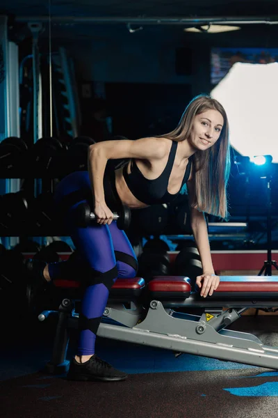 Vista lateral del hermoso entrenamiento de mujer fitness con pesas sobre banco de ejercicios en el gimnasio . — Foto de Stock