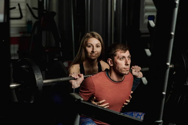 Active beautiful fitness model girl insists on squatting an athletic man in the gym.