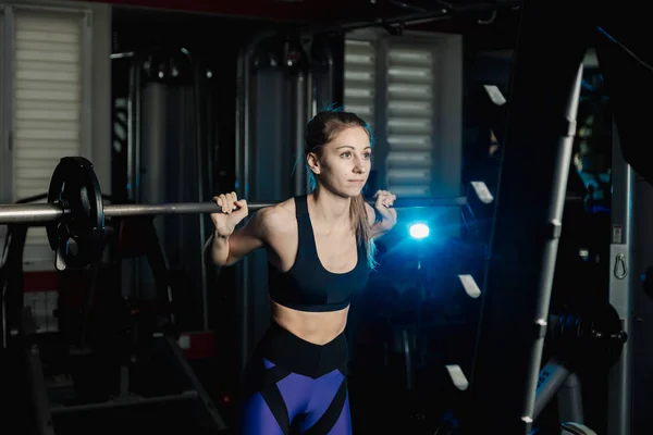 Active belle fille modèle de remise en forme s'accroupit avec un haltère sur les épaules dans la salle de gym . — Photo