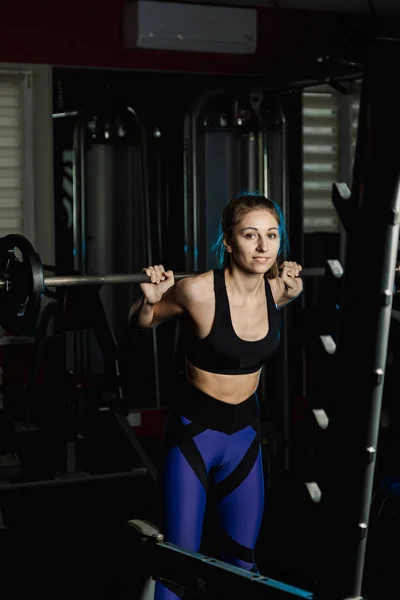 Activa hermosa chica modelo fitness se agacha con una barra en los hombros en el gimnasio . — Foto de Stock