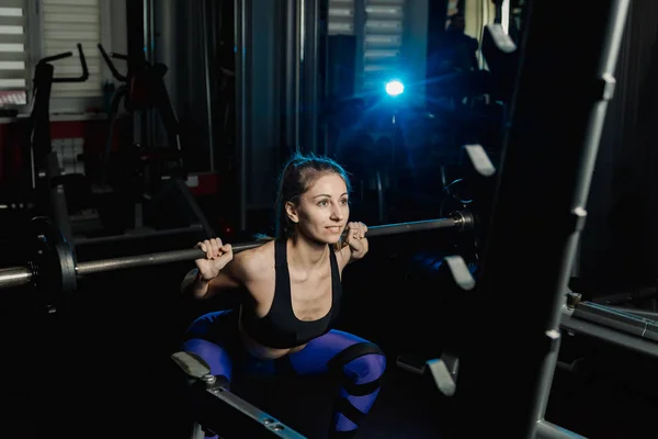 Activa hermosa chica modelo fitness se agacha con una barra en los hombros en el gimnasio . — Foto de Stock