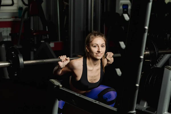 Active beautiful fitness model girl crouches with a barbell on the shoulders in the gym. — Stock Photo, Image