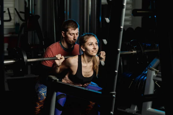 Handsome athletic man helps a beautiful girl to squat with a barbell on the shoulders in the gym.