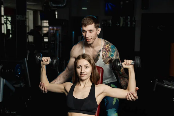 Active beautiful fitness model girl and her trainer training with dumbbells on the bench in the gym.