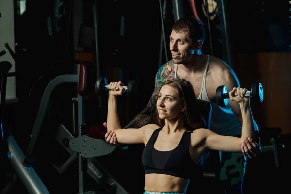 Chica activa modelo de fitness hermosa y su entrenamiento entrenador con pesas en el banco en el gimnasio . — Foto de Stock