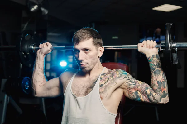 Yakışıklı atletik spor salonunda bankta oturmuş halter kaldırma omuz trenler.