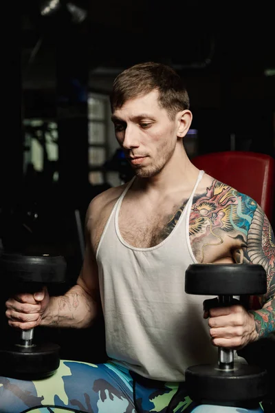 Spor salonunda bir bankta oturmuş dumbbells ile omuzlarını eğitim yakışıklı atletik erkek.