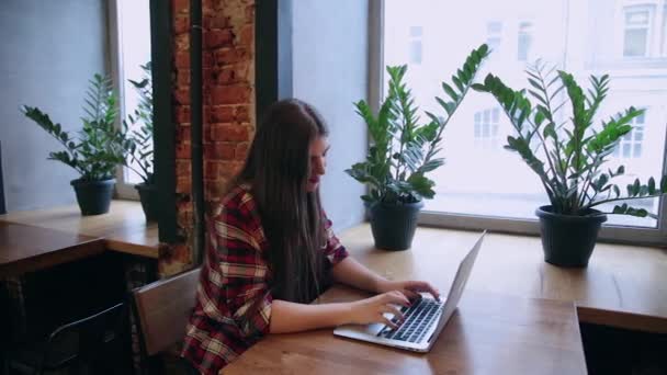 Una hermosa mujer de negocios está sentada en una mesa con un portátil en un café y trabajando . — Vídeo de stock