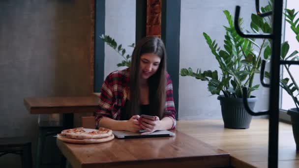 Mulher de negócios atraente bonita sentada com um telefone e um laptop em uma mesa em um café. Pizza na mesa . — Vídeo de Stock