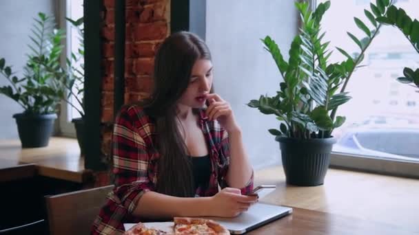 Vackra attraktiva affärskvinna sitter med en telefon och en bärbar dator vid ett bord på ett café. Pizza på bordet. — Stockvideo