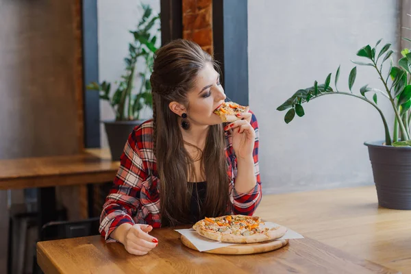 Hermosa y sexy chica sentada junto a la ventana en un café y comiendo pizza . — Foto de Stock