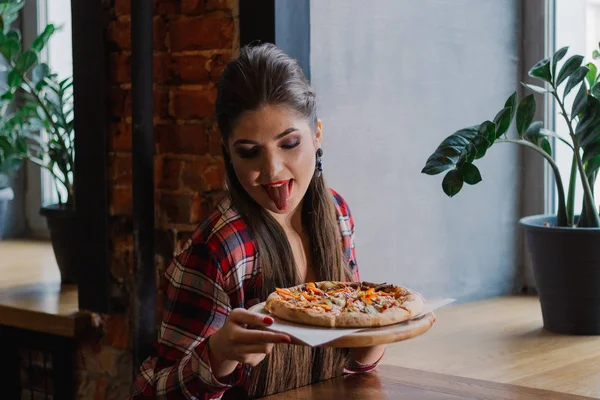 Hermosa y sexy chica sentada junto a la ventana en un café y sosteniendo una pizza . — Foto de Stock