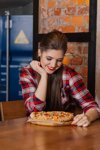 Beautiful and sexy girl sitting by the window in a cafe and eating pizza. — Stock Photo, Image