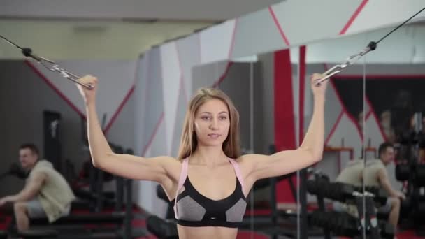 Belle jeune fille sportive entraîne biceps sur un simulateur de bloc dans la salle de gym . — Video