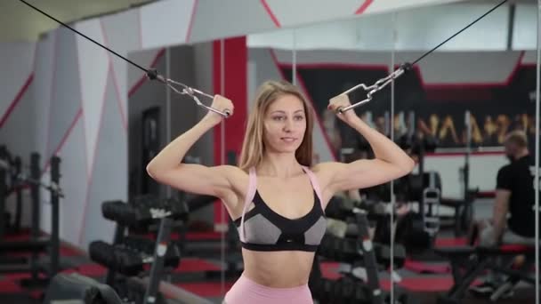 Hermosa chica atlética entrena bíceps en un simulador de bloque en el gimnasio . — Vídeos de Stock