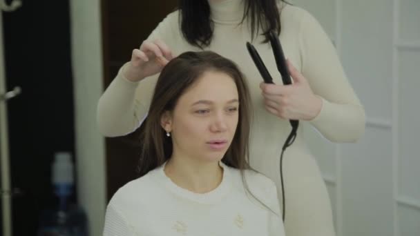 Mooie vrouw met bruin haar heeft het behandeld met een krultang door een kapper. Handheld real-time schot tot oprichting van. — Stockvideo