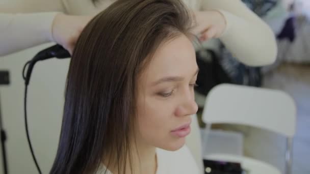 Красивая женщина с каштановыми волосами лечится щипцами для завивки волос парикмахером. Портативная съемка в режиме реального времени . — стоковое видео
