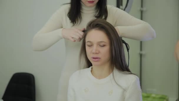 Красивая женщина с каштановыми волосами лечится щипцами для завивки волос парикмахером. Портативная съемка в режиме реального времени . — стоковое видео