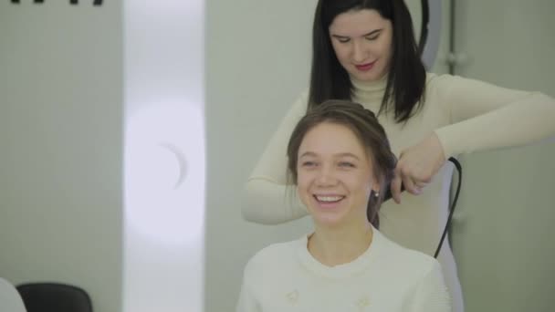 Hermosa mujer con cabello castaño está teniendo que tratarlo con una rizadora por un peluquero. De mano en tiempo real establecer tiro . — Vídeo de stock