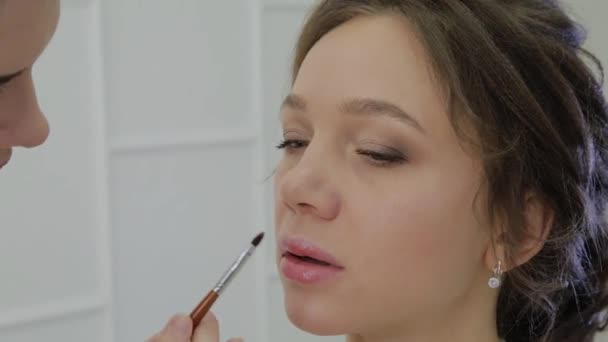 Καλλιτέχνης μακιγιάζ χρησιμοποιώντας πινέλο για εφαρμογή κραγιόν για τα χείλη όμορφη γυναίκα. Κοντινό πλάνο εφαρμογής λιπ γκλος χειλιών μόδα μοντέλο. Visagiste κάνει τα χείλη μακιγιάζ. Έννοια της ομορφιάς. — Αρχείο Βίντεο