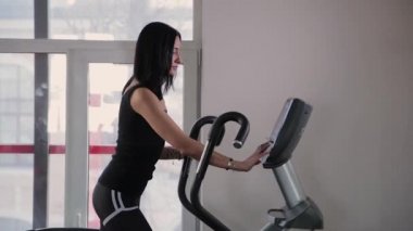 Güzel atletik genç kız spor salonunda eliptik bir eğitmen ayarlarını yapar.