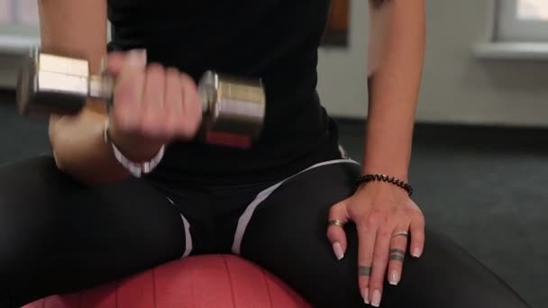 Joven mujer deportiva sentada en la pelota en forma y levantando pesas mientras entrena bíceps en el gimnasio . — Vídeo de stock