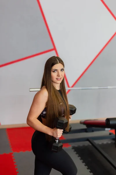 Muy hermosa chica entrena bíceps con mancuernas en el gimnasio . — Foto de Stock