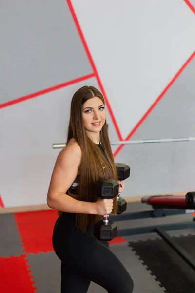 Muy hermosa chica entrena bíceps con mancuernas en el gimnasio . — Foto de Stock