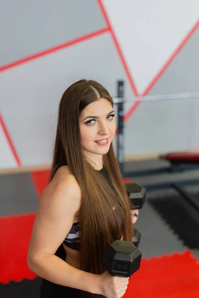 Muy hermosa chica entrena bíceps con mancuernas en el gimnasio . — Foto de Stock