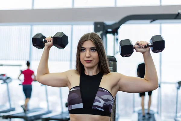 Very beautiful girl trains shoulders with dumbbells on the bench in the gym. — Stock Photo, Image